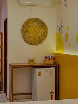 A room with a small white refrigerator, a wooden table with a basket and a glass, and a wall with a circular woven decoration and a simple floral drawing.