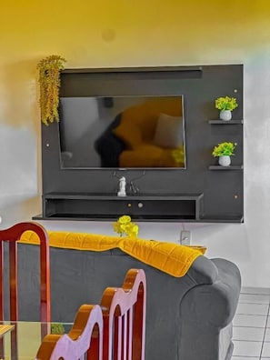 A living room with a flat-screen TV mounted on the wall, a grey sofa, a wooden chair, and a small table with decorative items.