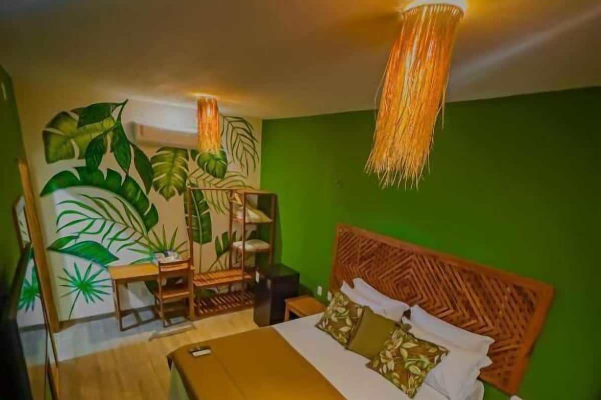 A bedroom with a bed, bedside tables, a chair, a wall with a plant mural, and a hanging light fixture.