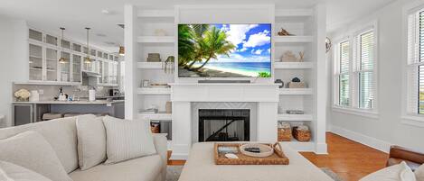 A living room with a TV, white furniture, a fireplace, and a kitchen in the background.