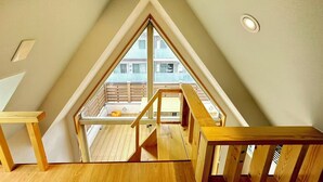 Un dormitorio en ático con techos altos, un gran ventanal triangular, vigas de madera y vistas a un balcón exterior.