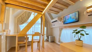 Una habitación con techo inclinado, vigas de madera, un televisor de pantalla plana, una planta en maceta y un comedor con sillas y una mesa.