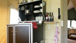 Una kitchenette compacta con cafetera, estantes con tazas y botellas, y un cubo de basura.