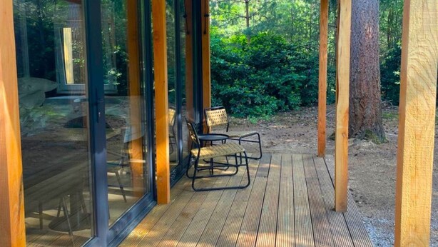 A covered wooden deck with glass doors leading to an outdoor seating area. - Outdoors Holten - Forest Cabin (Holten)