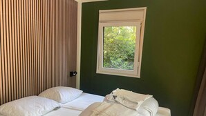 A double bed with white linens, a window with a view of greenery, and a wall with vertical wooden slats. - Outdoors Holten - Forest Cabin (Holten)