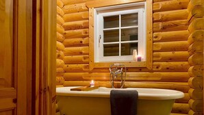 Une salle de bain rustique, aménagée dans une cabane en rondins, avec une baignoire sur pieds, une fenêtre et un sol carrelé.