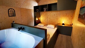 Una camera da letto con una vasca da bagno ampia, un letto con cuscini, un comodino con una lampada e uno scaffale con oggetti vari.