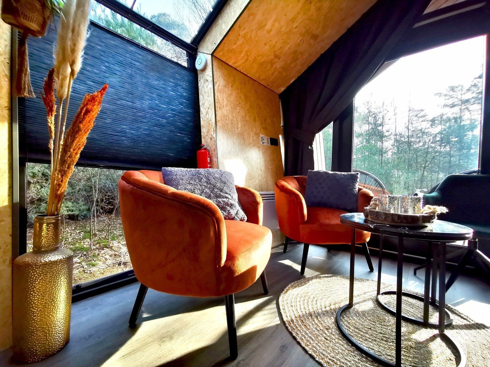 A modern interior with a large window, two orange chairs, a round table, and a vase with dried plants.