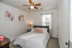 Une chambre à coucher comprenant un lit, un ventilateur de plafond, une table de chevet avec une lampe, et deux tableaux abstraits accrochés au mur.