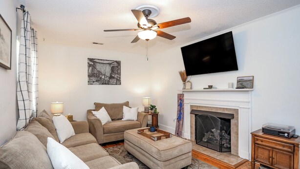 A living room with a sofa, ottoman, fireplace, television, and ceiling fan. - Parkins Mill Bungalow (Greenville)