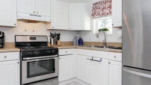 A kitchen with white cabinets, stainless steel appliances, a stove, oven, sink, and a window with a floral curtain. - Parkins Mill Bungalow (Greenville)