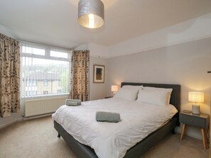 Quarto com cama, abajures, janela com cortinas, radiador e um quadro na parede.