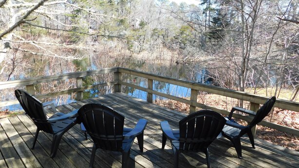 A wooden deck with four chairs and a table, overlooking a body of water and trees. - Iconic 70s style cute condo cottage in Deck 2, on a pond! (CS0055) (Wellfleet)