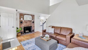 Een woonkamer met een open haard, een bruine leren bank, een grijze poef en een houten bijzettafeltje.