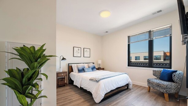 A bedroom with a bed, bedside tables, a lamp, a plant, a window with blinds, and a view of buildings outside. - Peaceful ! Free Parking ! 30 min to MetLife ! (City of Orange)