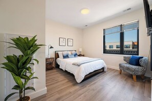 A bedroom with a bed, bedside tables, a lamp, a plant, a window with blinds, and a view of buildings outside. - Peaceful ! Free Parking ! 30 min to MetLife ! (City of Orange)