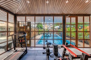 A gym with exercise equipment, a treadmill, and a view of an outdoor swimming pool through large windows. - Elegant 1bhk in Candolim Pool Gym Near Beach (Candolim)