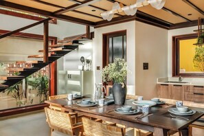 A modern kitchen with a dining area, featuring a staircase, wooden furniture, and a vase with flowers. - Casa Melina - 1 5BR Private Beach House in Anjuna (Anjuna)