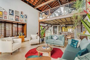 A spacious living area with a high ceiling, a staircase leading to an upper level, and a kitchen visible through a glass partition. - Casa Melina - 1 5BR Private Beach House in Anjuna (Anjuna)