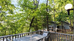Un patio in legno con tavolo e sedie, immerso nel verde tra alberi e con una casa sullo sfondo.