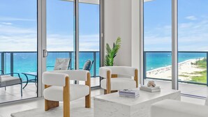 Habitació moderna amb una gran finestra que dona a un balcó amb vista al mar i a la platja. Hi ha dues cadires blanques amb coixins i una taula petita amb un llibre i un objecte decoratiu.