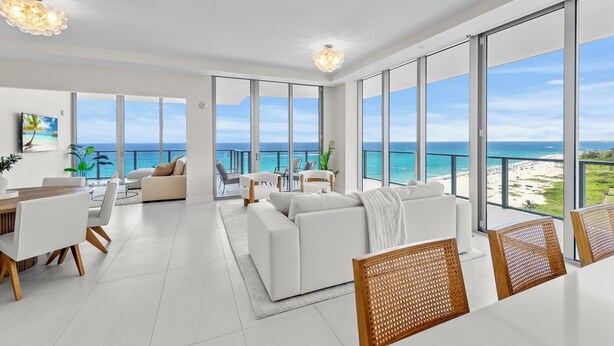Un ampli sal ó amb una finestra gran amb vista a una platja, una zona de dinar amb taula de fusta i un sofà blanc.