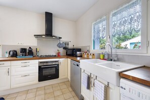 A kitchen with a stove, oven, dishwasher, and a window with lace curtains. - Le Cardoue: Peaceful Gîte Between Land and Sea in the Marais Breton Vendéen (Saint-Gervais)