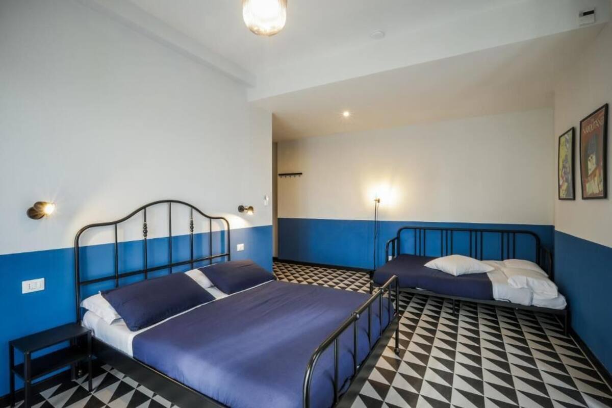 A room with two beds, each with a metal frame and blue bedding. There are wall-mounted lights and a checkered floor pattern.