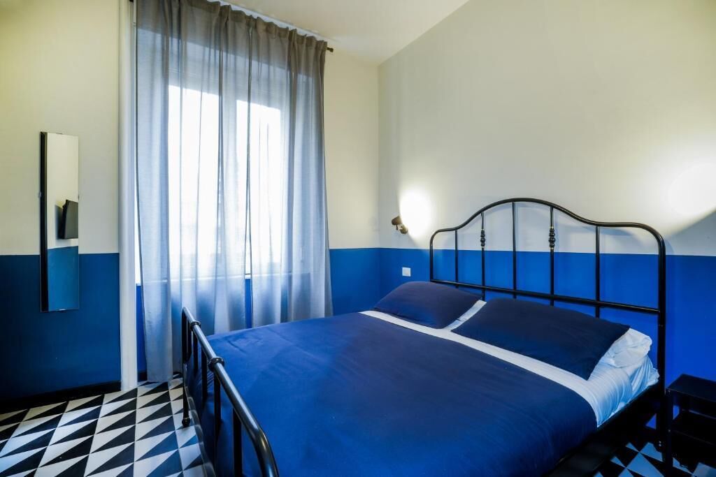 A room with a bed, a mirror, a window with curtains, and a geometric-patterned floor.