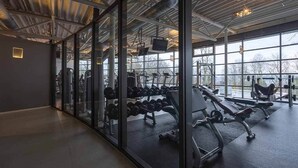 Ein Fitnessstudio mit Trainingsgeräten, großen Fenstern und einer modernen, industriell gestalteten Decke.