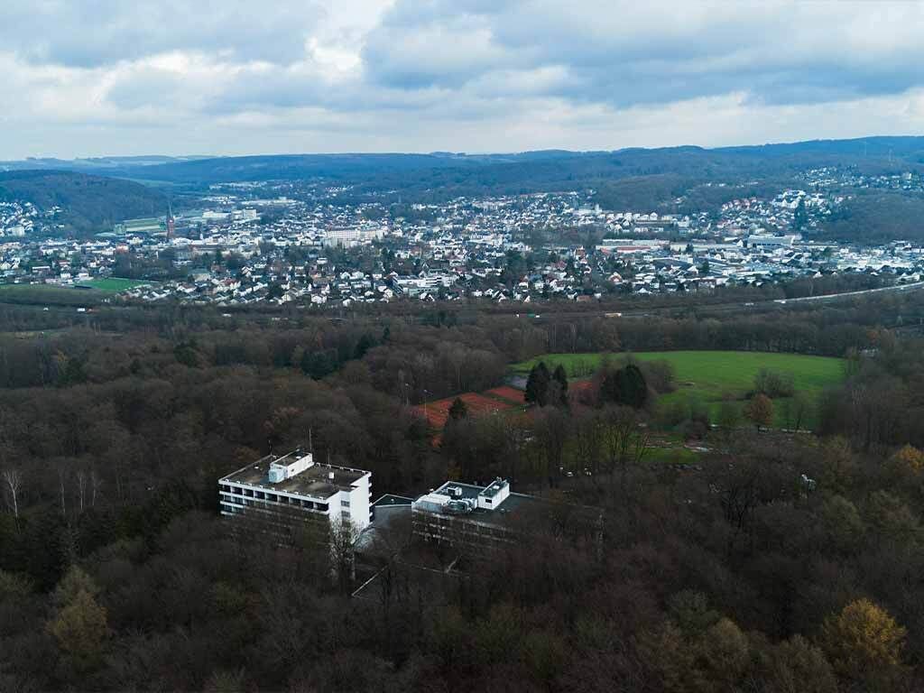 Eine Vogelperspektive auf eine Stadt mit einem markanten Gebäude im Vordergrund, umgeben von dichtem Wald und einer Mischung aus Wohn- und Geschäftsgebäuden im Hintergrund.