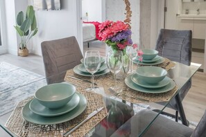 Ein Esstisch ist gedeckt mit grünen Tellern und Gläsern, eine Blumenvase steht darauf, und im Hintergrund ist ein modernes Interieur mit einer Pflanze und einem Bett zu sehen.