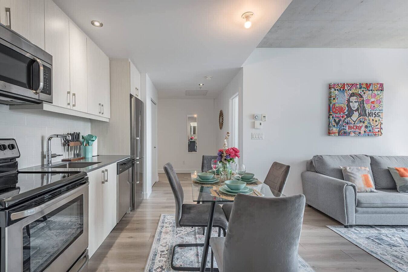 A modern kitchen with stainless steel appliances, a dining area with a table set for four, and a grey sofa in the living room.
