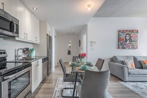 Eine moderne Küche mit Edelstahlgeräten, ein Essbereich mit Tisch für vier Personen und ein graues Sofa im Wohnzimmer.