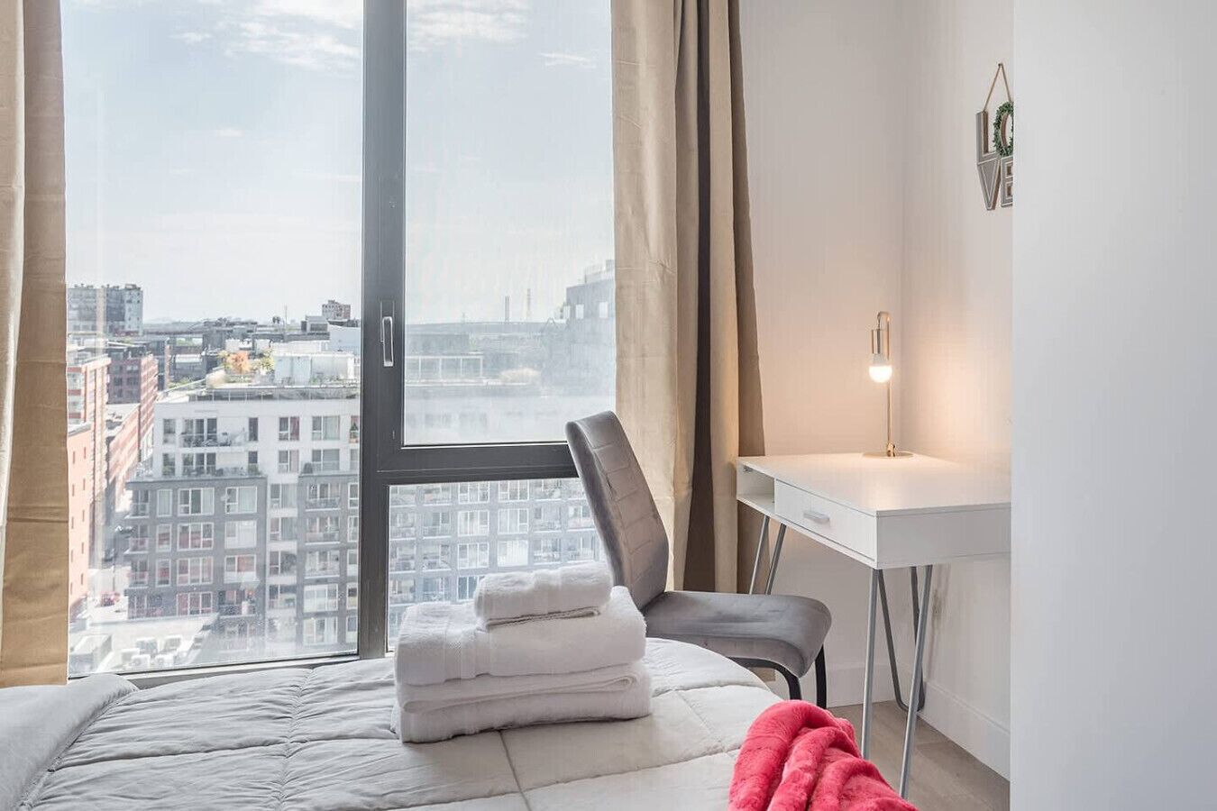 A bedroom with a large window, a bed, a chair, a desk, and a lamp.