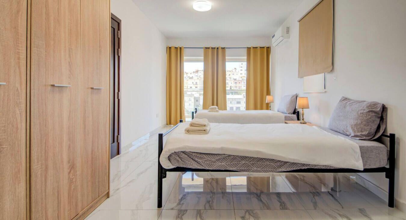 A hotel room with a bed, bedside tables, a wardrobe, and a view of buildings through the window.