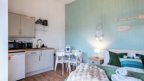 Ett kompakt hotellrum med ett litet pentry, en säng med ett grönt och vitt randigt överkast, en drömfångare och en träpaddel på väggen.