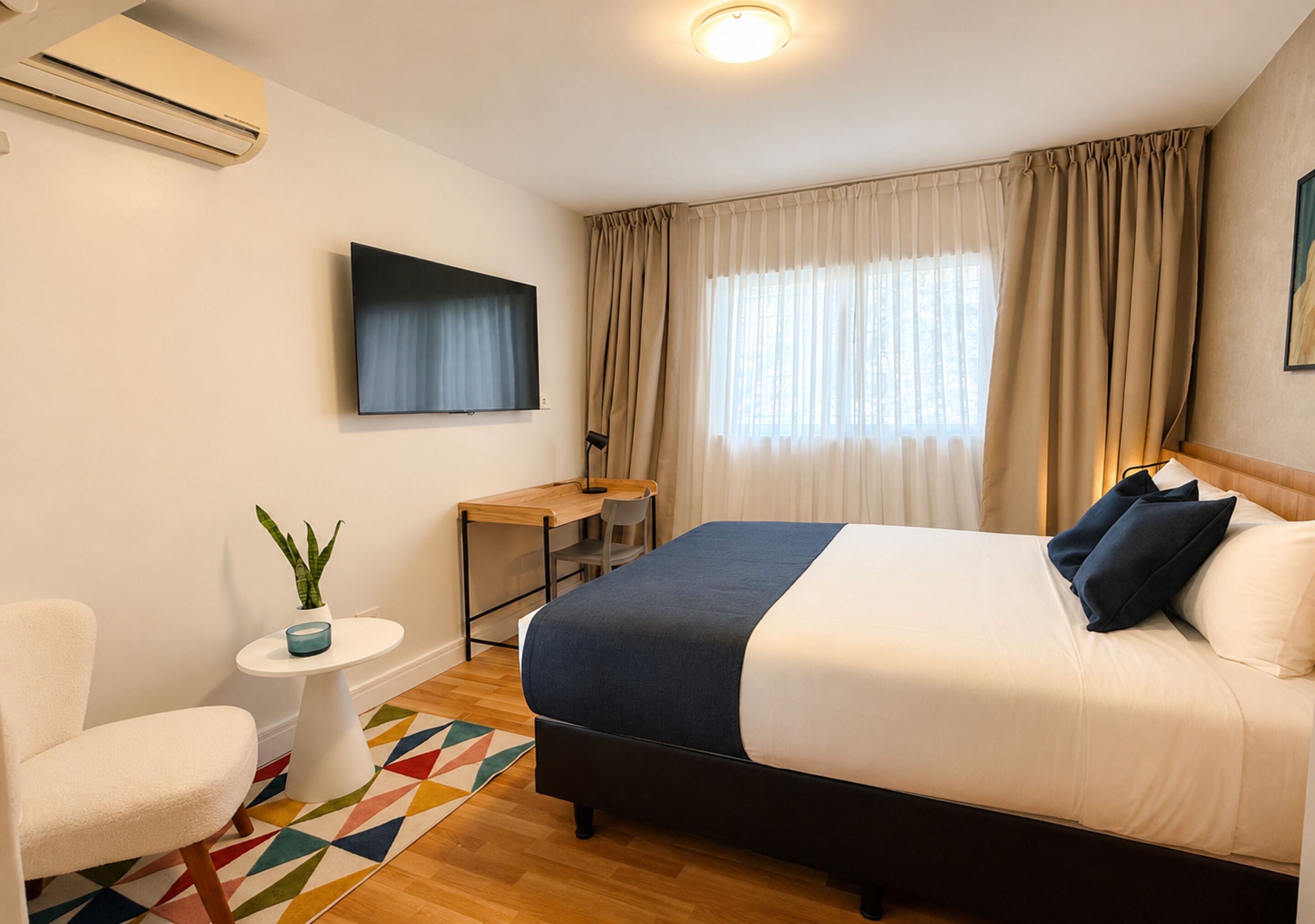 A hotel room with a bed, a desk, a chair, a small table with a plant, and a colorful rug.