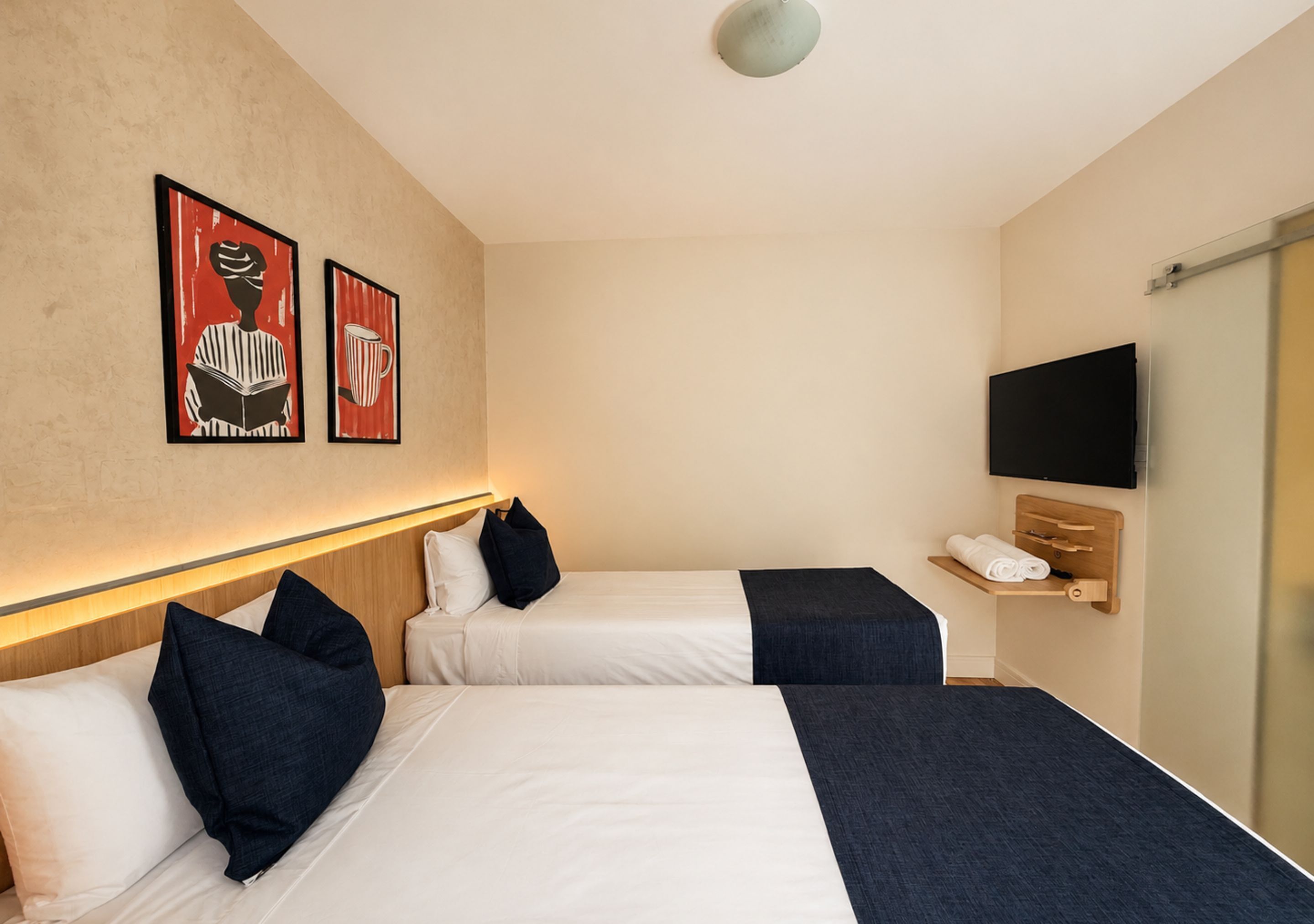 Ein Hotelzimmer mit zwei Betten, einem Fernseher, einem Regal mit Handtüchern und Bildern an der Wand.