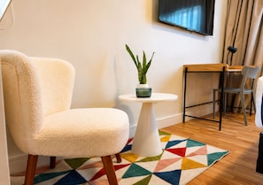 Une pièce avec un fauteuil moelleux, une petite table sur laquelle est posée une plante, un tapis coloré et un téléviseur fixé au mur.