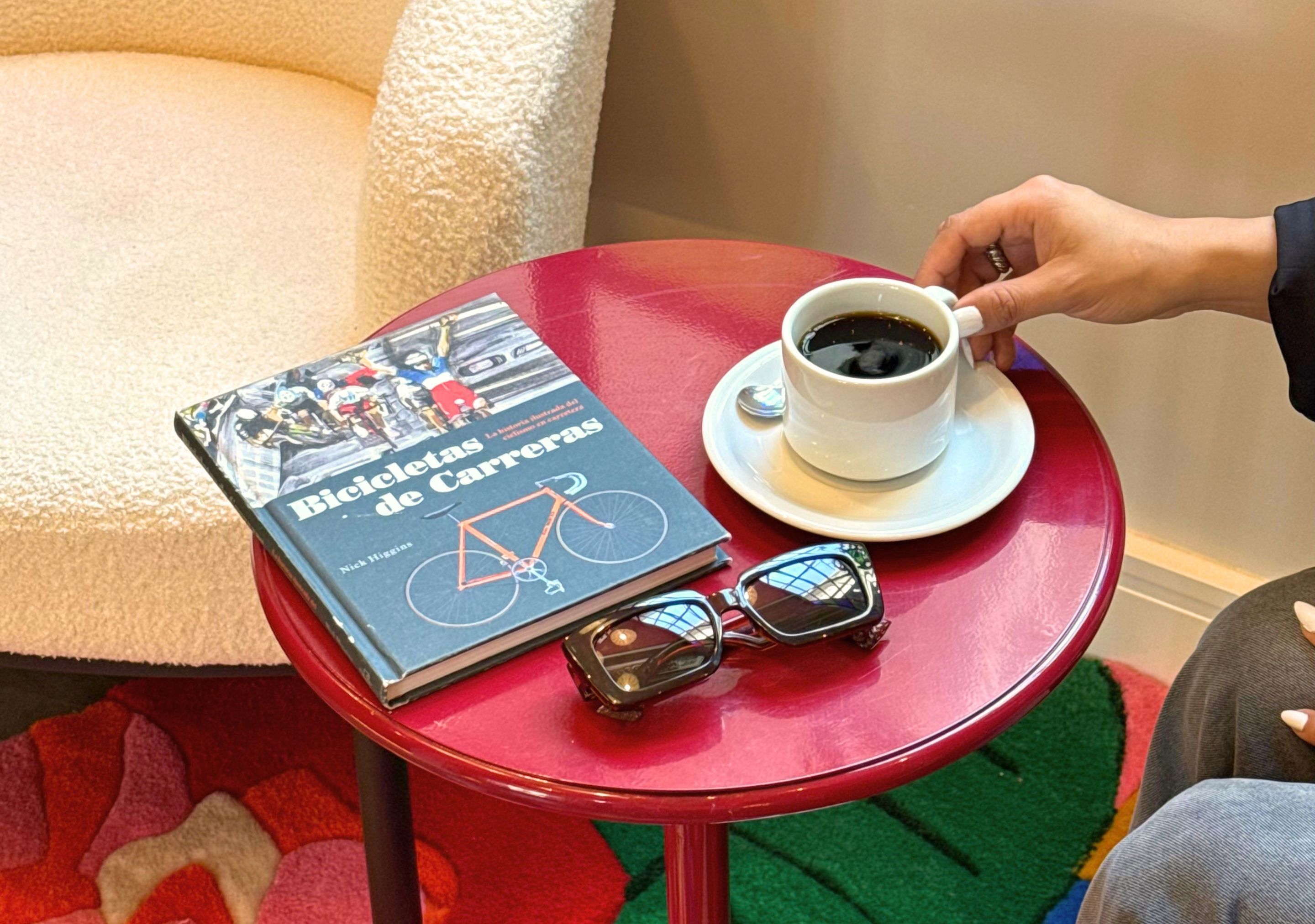 A book titled 'Bicicletas de Carrera' on a red table with a cup of coffee and sunglasses.
