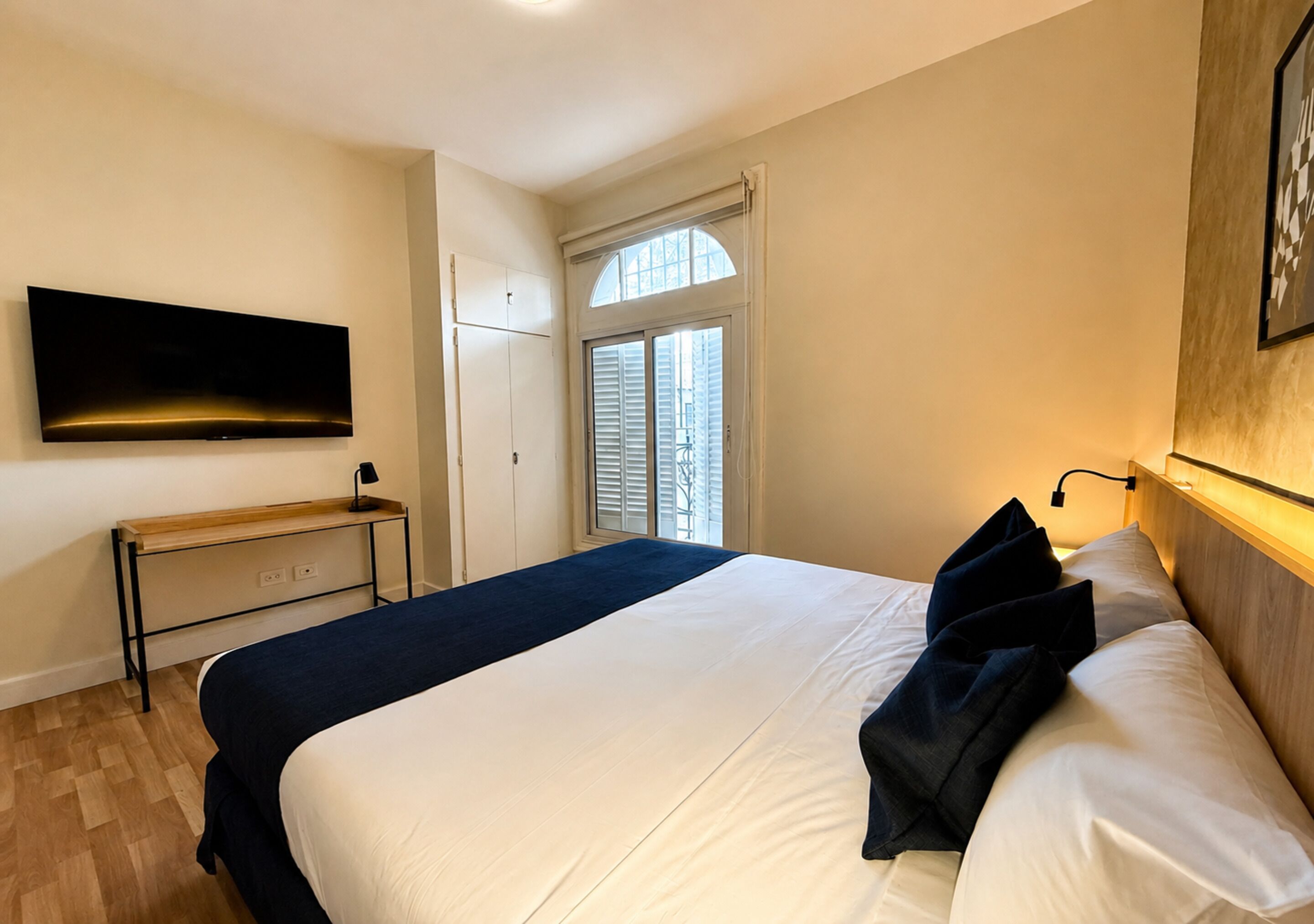 Una habitación de hotel con cama, televisor en la pared, escritorio y ventana con contraventanas.
