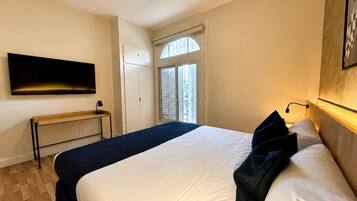 Una habitación de hotel con cama, televisor en la pared, escritorio y ventana con contraventanas.