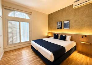 A bedroom with a large bed, wooden flooring, a window with shutters, wall-mounted air conditioning, and two framed pictures.