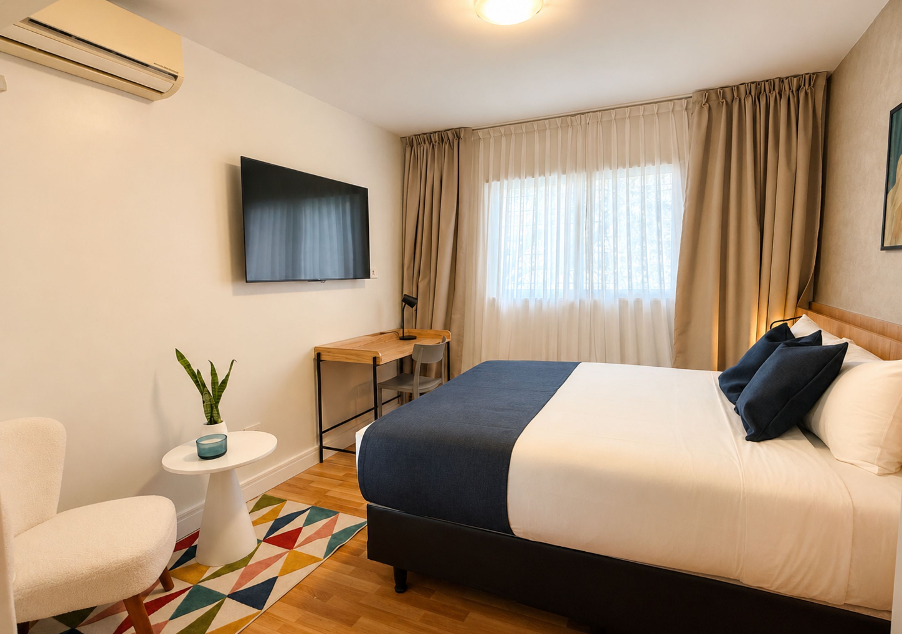 Habitación de hotel con cama, escritorio, silla, una mesita con planta y alfombra colorida.