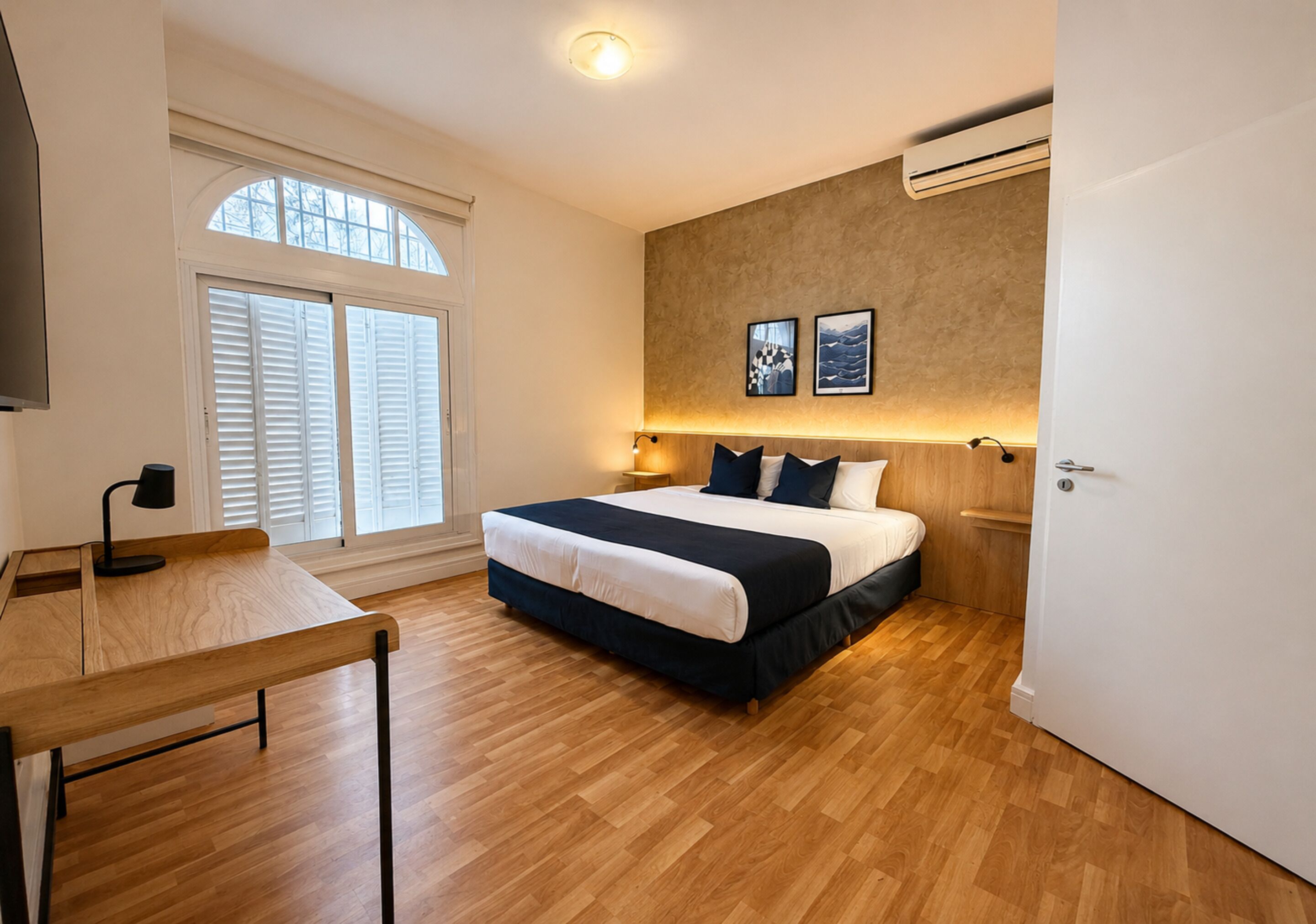 A bedroom with a bed, a desk, a window with shutters, and wall-mounted artwork.