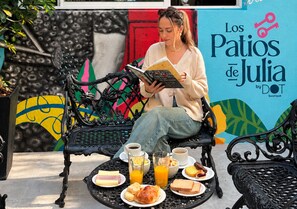 A person seated on a bench with a book, surrounded by a table with breakfast items and a wall with colorful artwork.