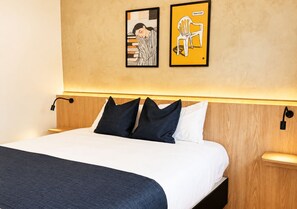 Una cama bien tendida con ropa de cama blanca y azul oscuro, un cabecero de madera y dos obras de arte en la pared que muestran a una persona durmiendo y una silla con un gato.