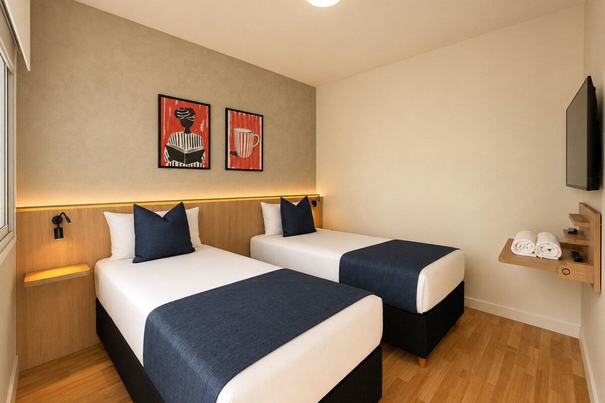 A hotel room with two beds, a wooden floor, a shelf with towels, and two framed pictures on the wall.