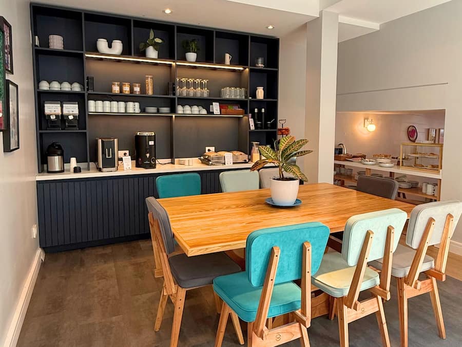 Un espace repas moderne avec une table et des chaises en bois, une cuisine intégrée dotée d’étagères et une plante en pot.